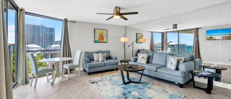 A modern living room with a sofa, coffee table, and dining area, large windows with curtains, and a ceiling fan.