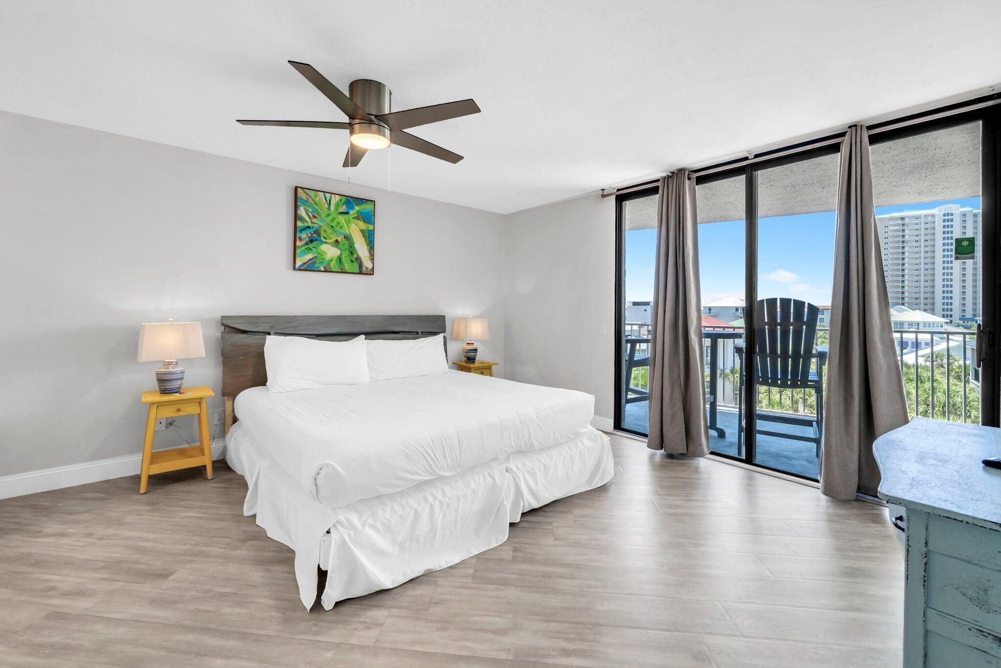 A modern hotel room with a large bed, a ceiling fan, a painting, and a balcony with a view of buildings.