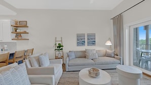 A modern living room with a sofa, two armchairs, a coffee table, and a dining area with a table and chairs. There are wall-mounted shelves with decorative items and a ladder. - Emerald by Alma (Santa Rosa Beach)