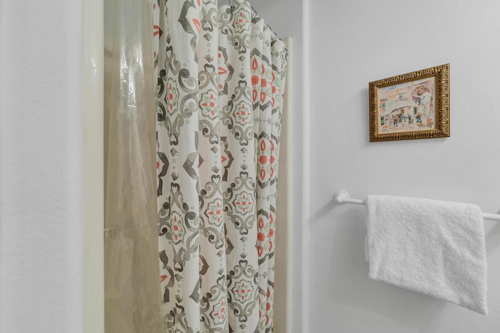 Een badkamer met een gordijn met een patroon, een handdoekhouder met een witte handdoek, en een ingelijst beeld op de muur.