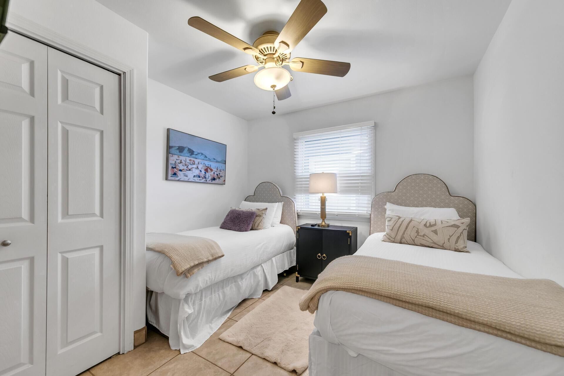 A bedroom with two beds, a ceiling fan, a painting, a nightstand, and a window with blinds.