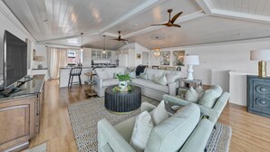 A spacious living area with a large sofa, a coffee table, and a TV. There is a kitchen in the background with white cabinets and a dining area. - Henderson Haus (Destin)