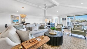A spacious living room with a large sofa, a coffee table, and a round side table. There is a ceiling fan, a chandelier, and a large window with blinds. - Henderson Haus (Destin)