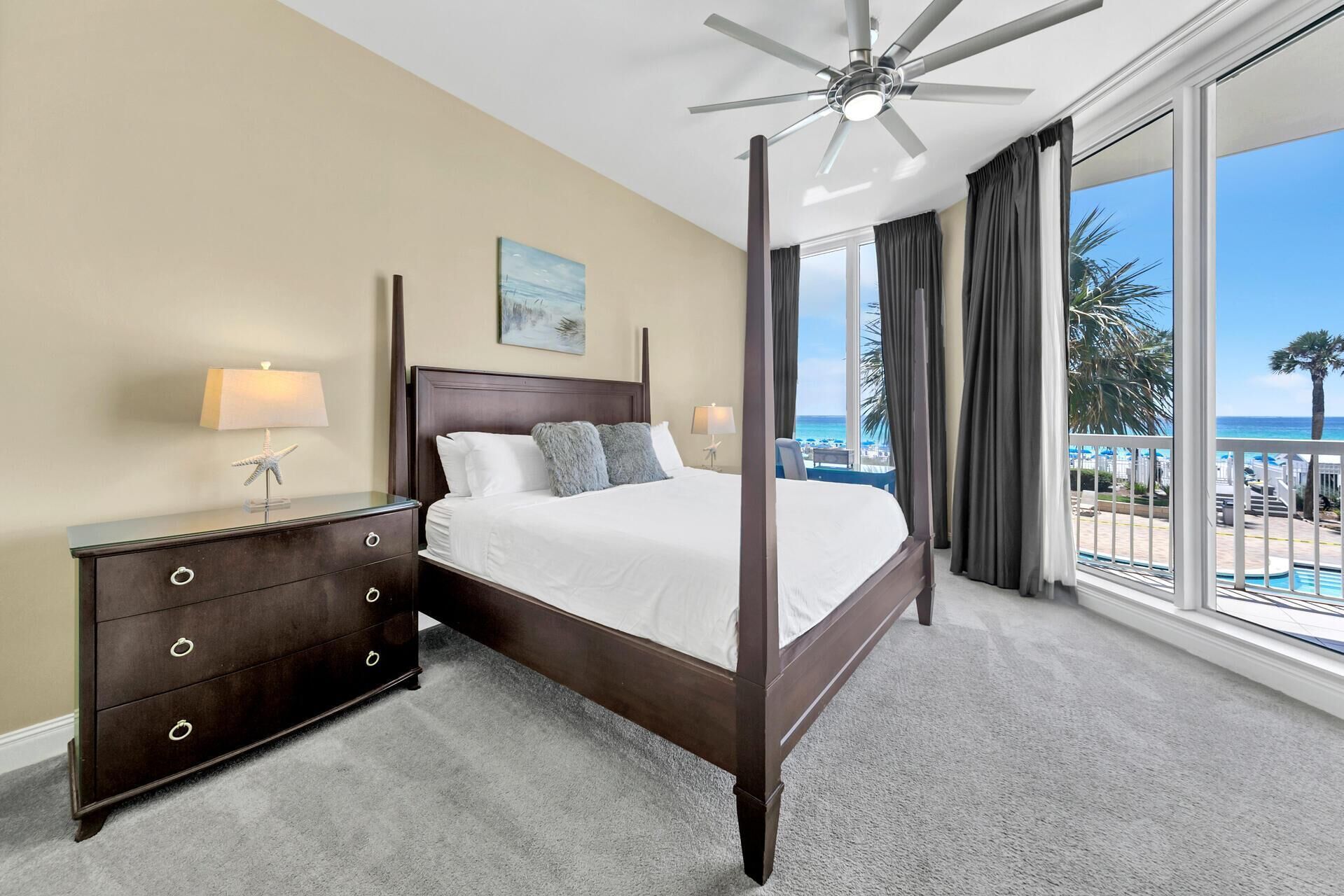 A four-poster bed with a view of the ocean, a ceiling fan, and a nightstand with a lamp.