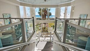 A modern hotel interior with a glass staircase leading to a balcony with a view of the beach and palm trees. - Silver Beach Towers - Penthouse106e (Destin)