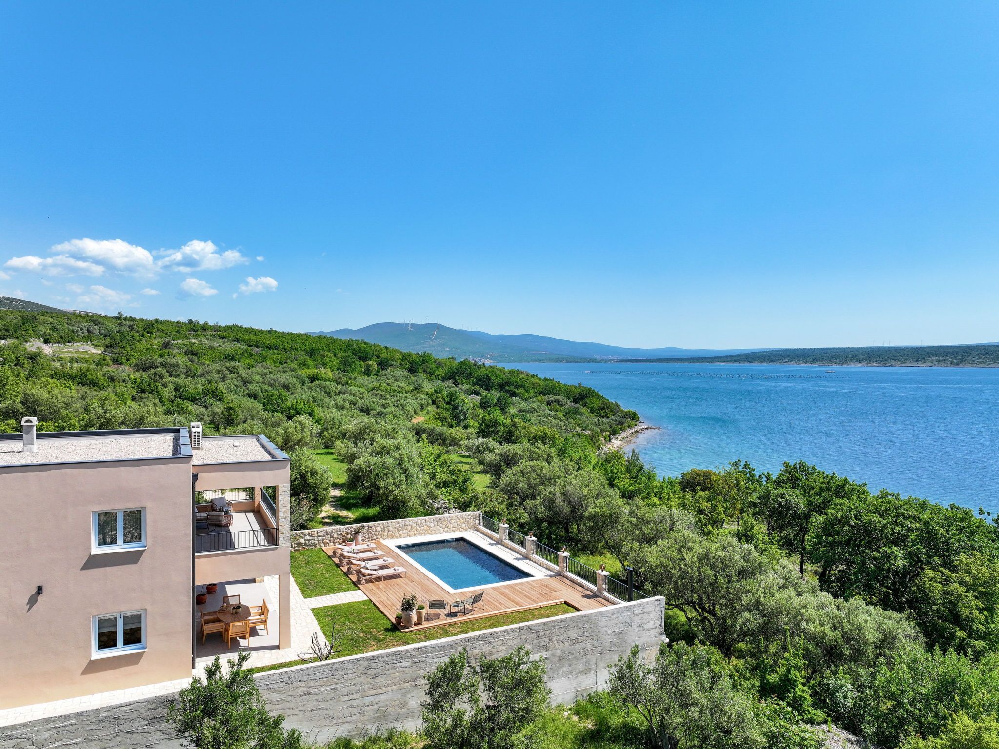 A modern two-story building with a swimming pool, outdoor seating, and a view of the sea.