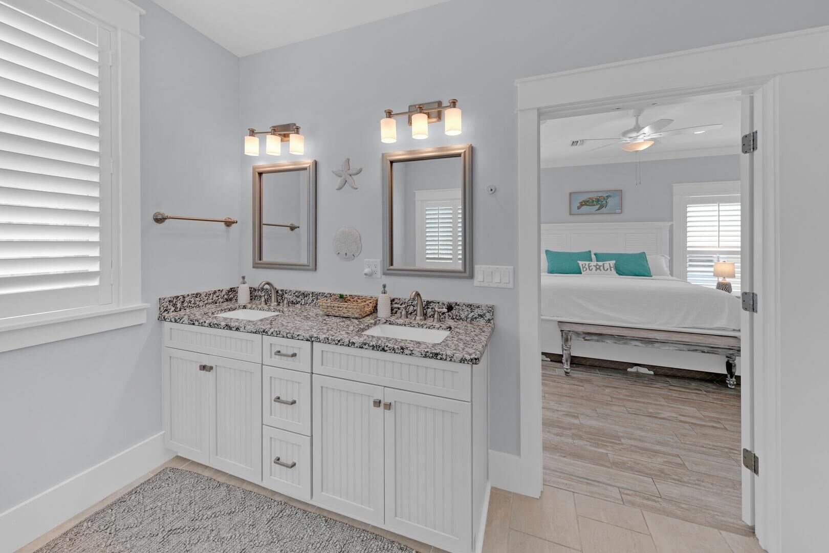 A bathroom with white cabinetry, a granite countertop, and a double vanity. There are two wall-mounted mirrors with light fixtures, a towel rack, and a starfish decoration. The adjacent bedroom has a bed with a white headboard, a teal pillow, and a painting on the wall.