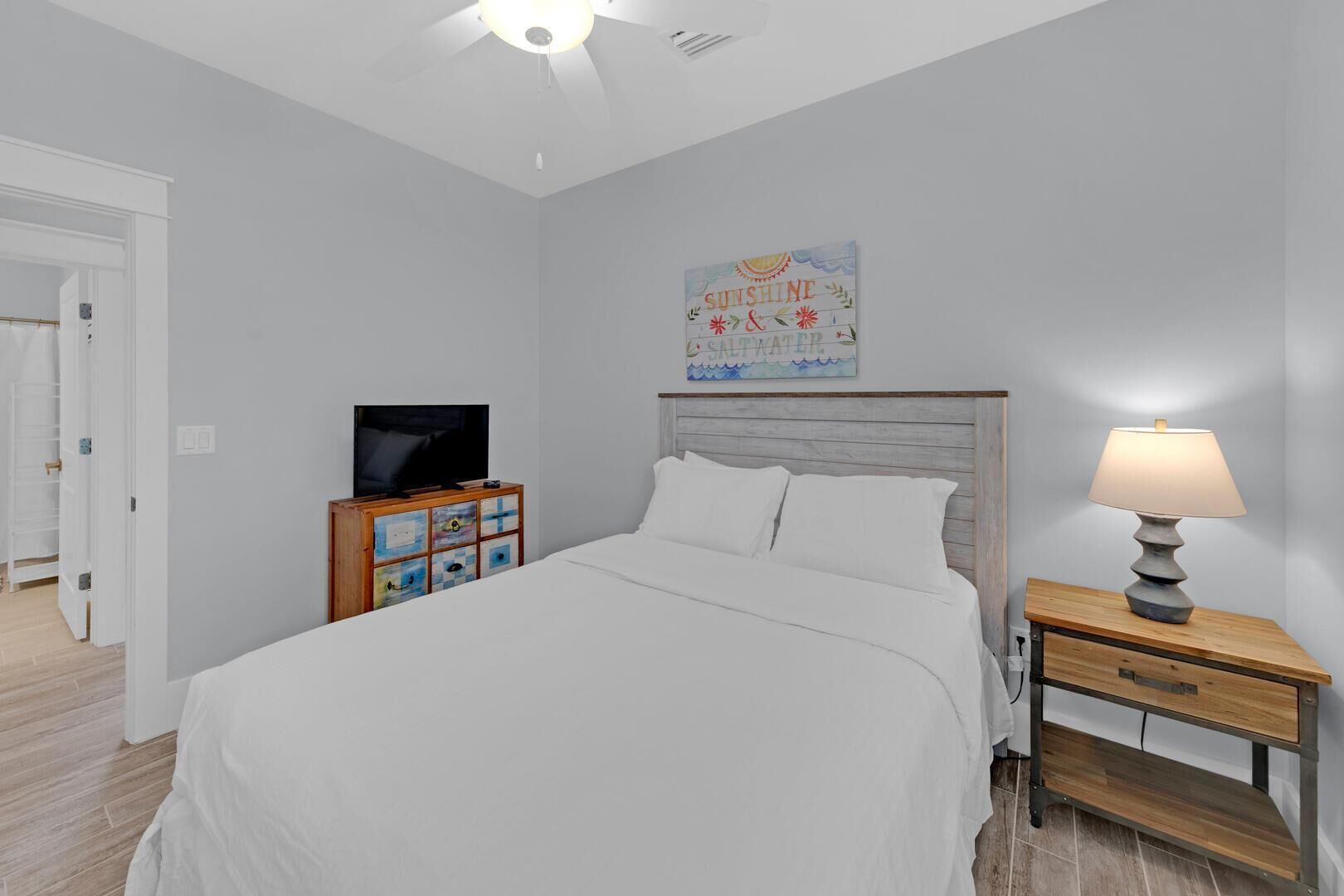 Une chambre à coucher avec un lit, un téléviseur posé sur une commode en bois, une lampe de chevet et une œuvre d’art murale.