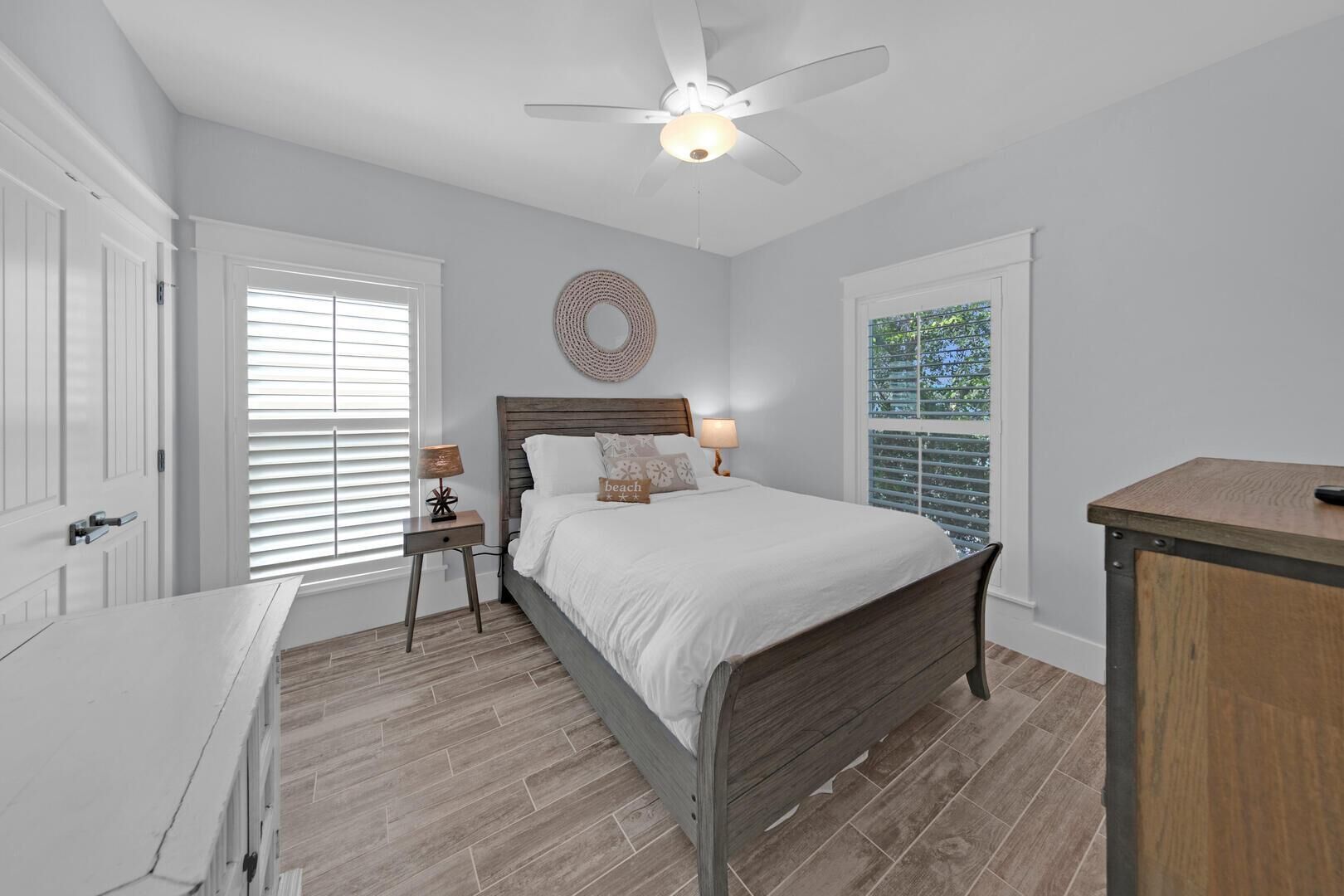 A bedroom with a wooden bed, a ceiling fan, a bedside table with lamps, and a view through a window.