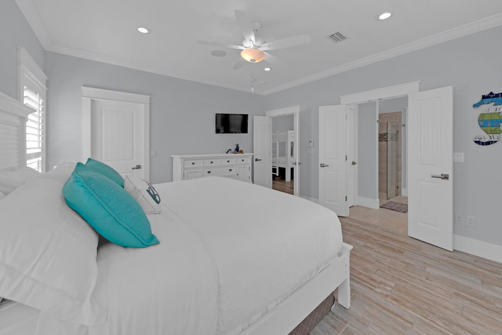 A bedroom with a large bed, a ceiling fan, a TV, and a bathroom visible through an open door.