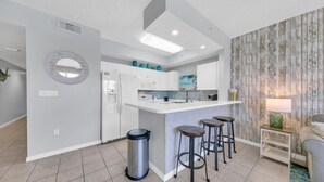 A modern kitchen with white cabinetry, a central island with stools, a stainless steel trash can, and a decorative mirror on the wall.