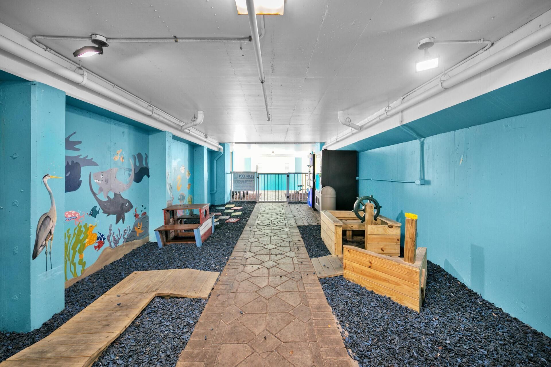 Un espace de jeux pour enfants agrémenté d’une fresque murale, de structures de jeux en bois et d’un chemin pavé de carreaux hexagonaux.