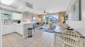 A modern kitchen with white cabinetry, a dining area with a table and chairs, and a view of the pool and building in the background.