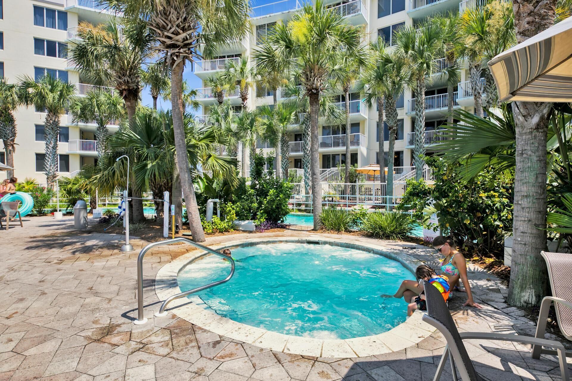 A pool area with a circular pool, palm trees, and a multi-story building with balconies.