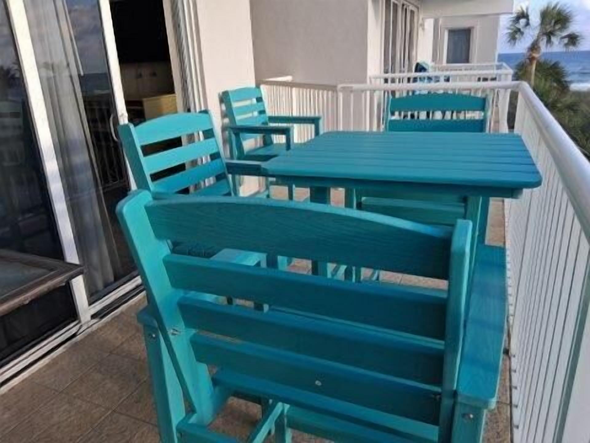 A balcony with turquoise chairs and a table, overlooking a beach.