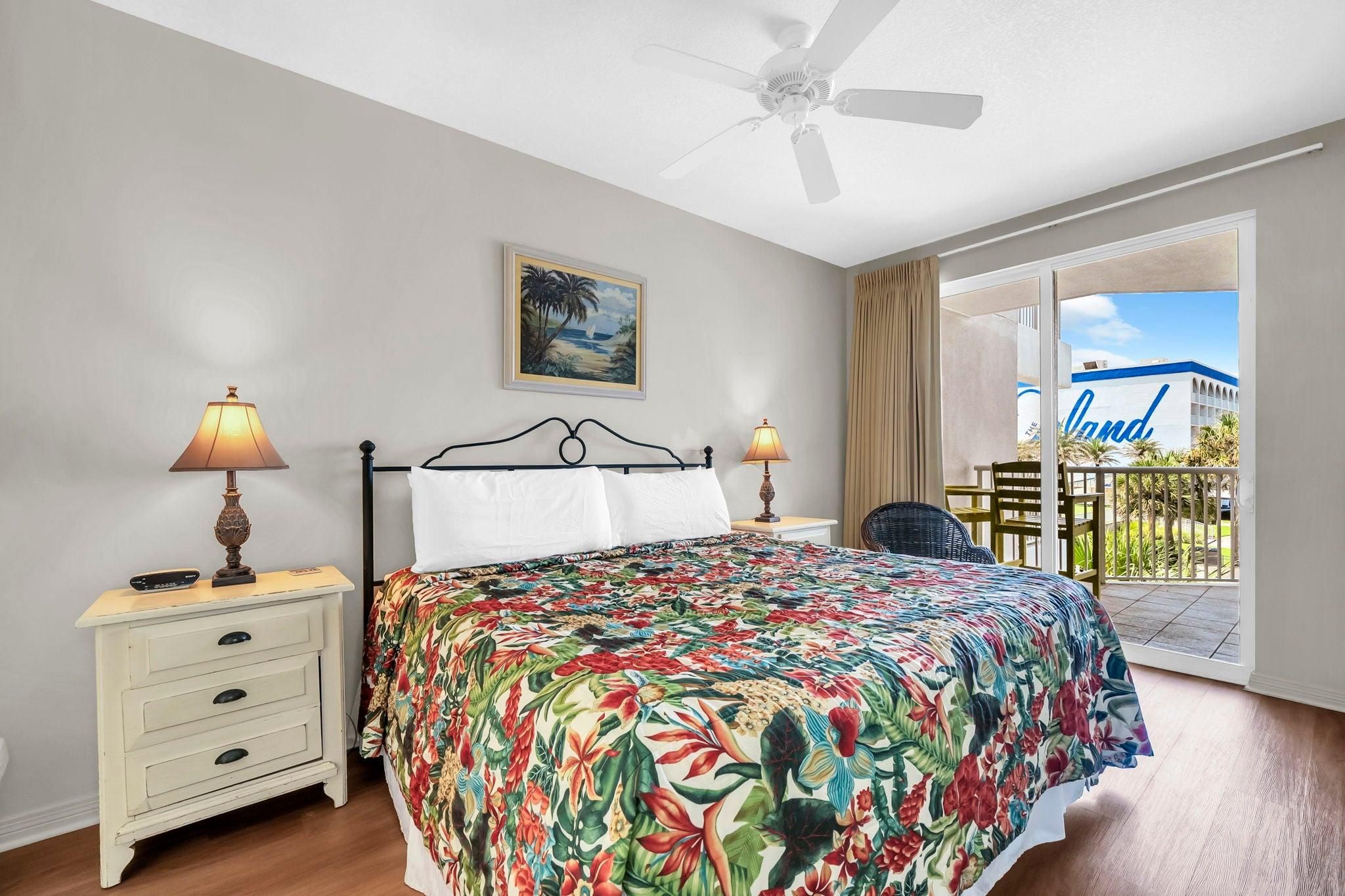 A bedroom with a bed, two bedside lamps, a nightstand, a ceiling fan, a painting, and a view of a building with 'The Land' written on it.