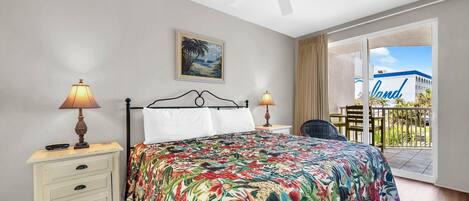 A bedroom with a bed, two bedside lamps, a nightstand, a ceiling fan, a painting, and a view of a building with 'The Land' written on it.