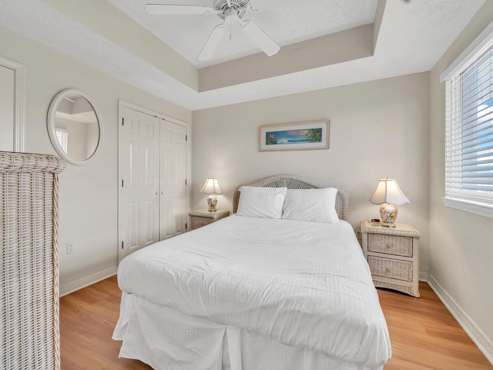 A bedroom with a large bed, two bedside tables with lamps, a wicker chair, a ceiling fan, a door, and a painting on the wall.