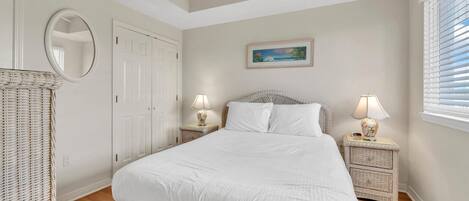 A bedroom with a large bed, two bedside tables with lamps, a wicker chair, a ceiling fan, a door, and a painting on the wall.