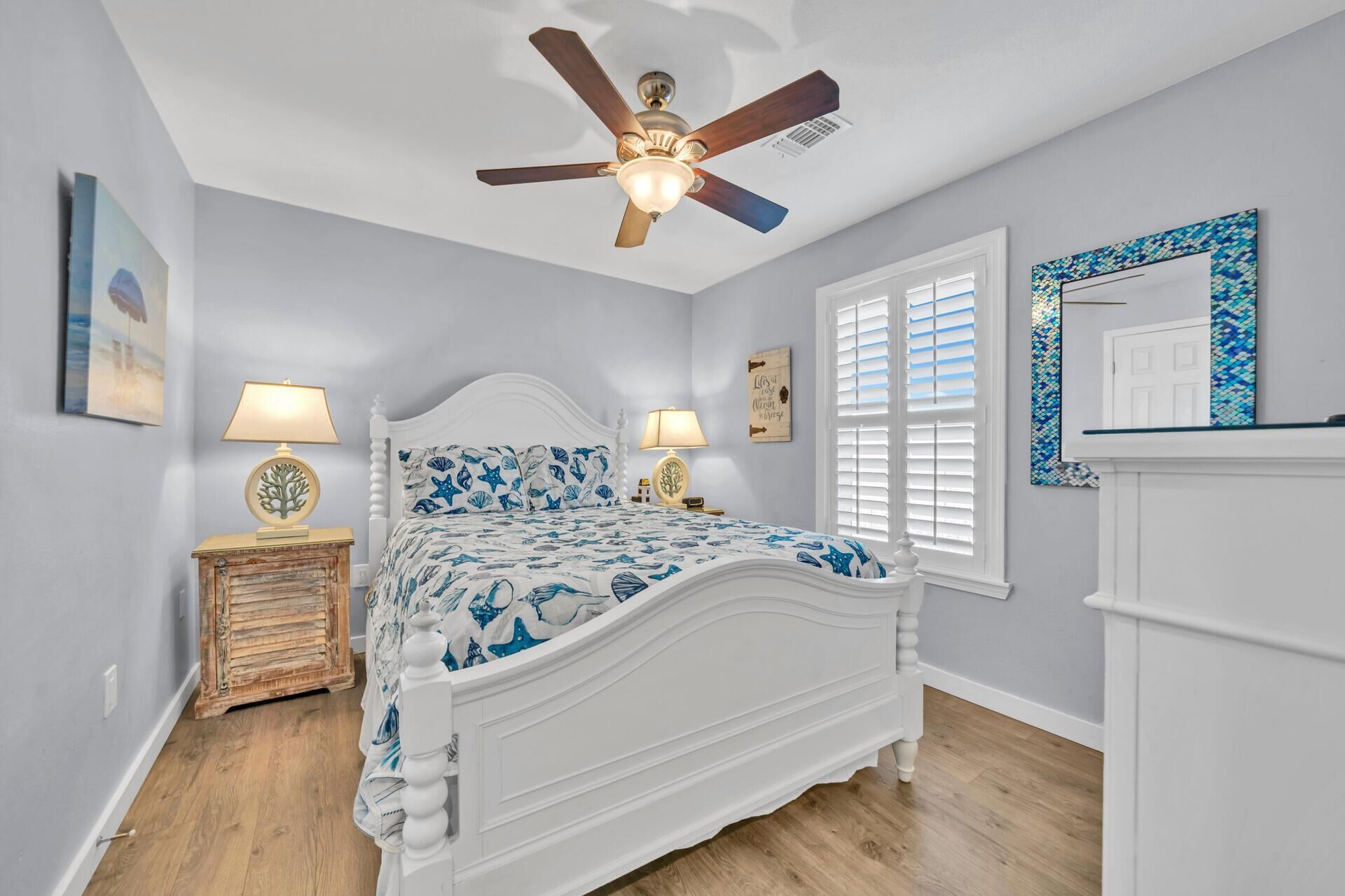 Une chambre à coucher avec un lit blanc, deux tables de chevet avec des lampes, un ventilateur de plafond, une fenêtre avec des stores et un miroir décoratif.