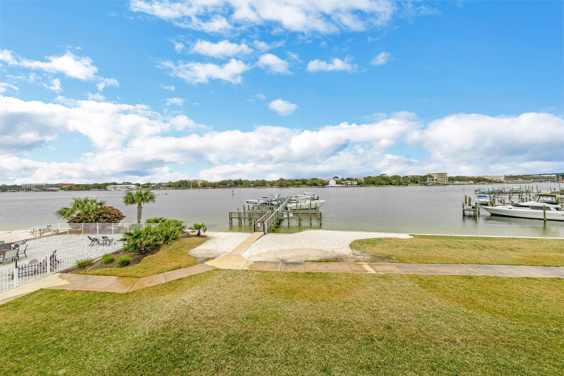 A marina with boats docked, a grassy area with a pathway, and a view of the water and distant buildings.