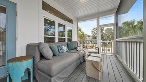 A covered porch with a gray sofa, teal side table, and a view of palm trees. - Cloud Nine (Santa Rosa Beach)