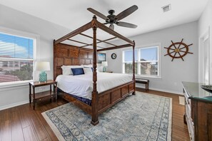 A four-poster canopy bed with a ceiling fan, a ship's wheel wall decoration, and a view of the city through large windows. - A Spyglass (Destin)