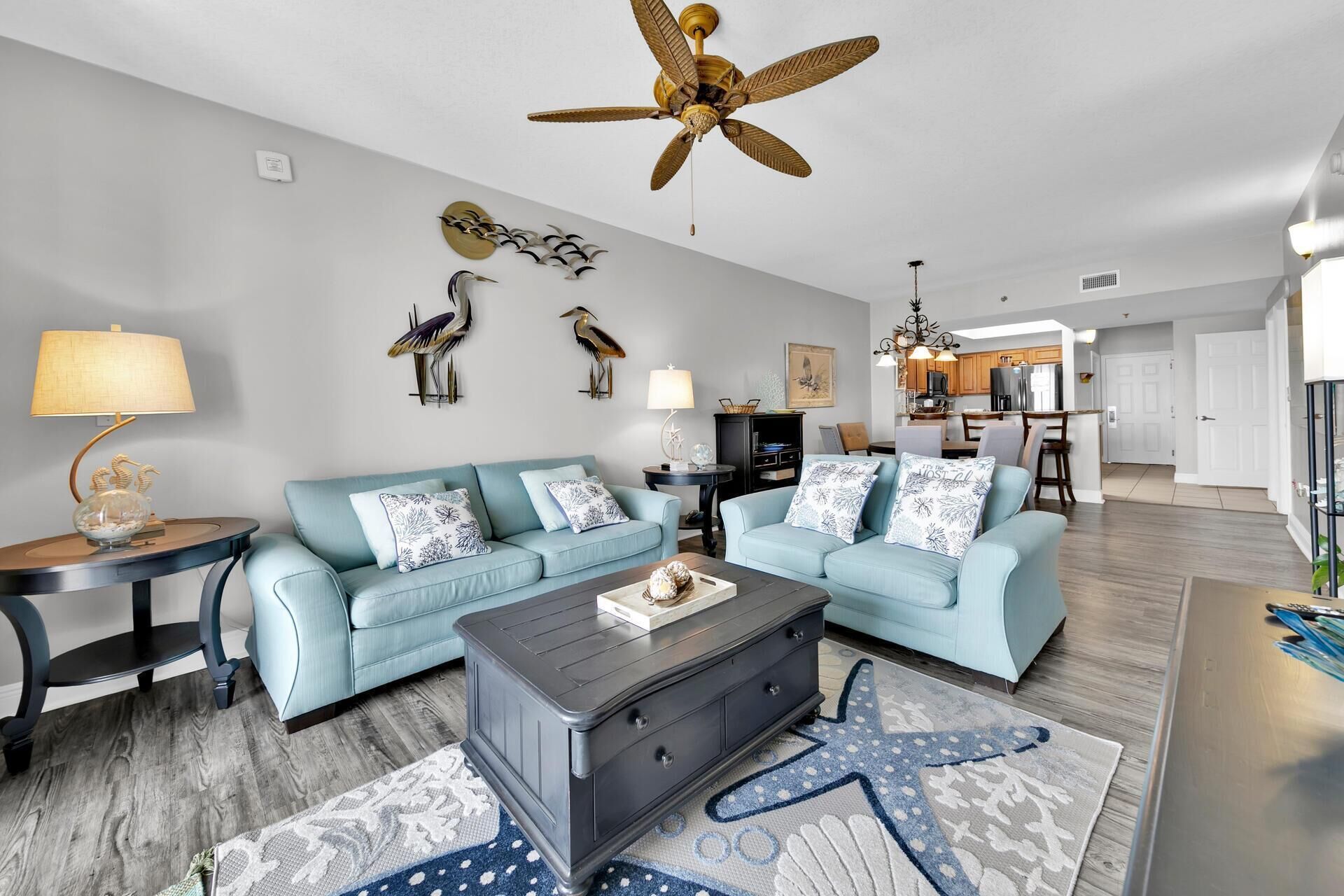 Ein Wohnzimmer mit blauem Sofagarnitur, einem Couchtisch und einem Teppich mit Seestern-Motiv. Man sieht einen Deckenventilator, Wandkunst und eine Küche im Hintergrund.