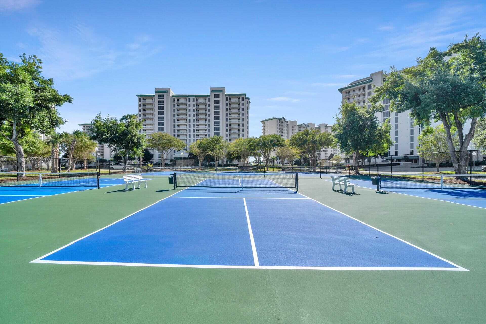 Un court de tennis en plein air, doté d’un filet, entouré d’arbres et de bâtiments.