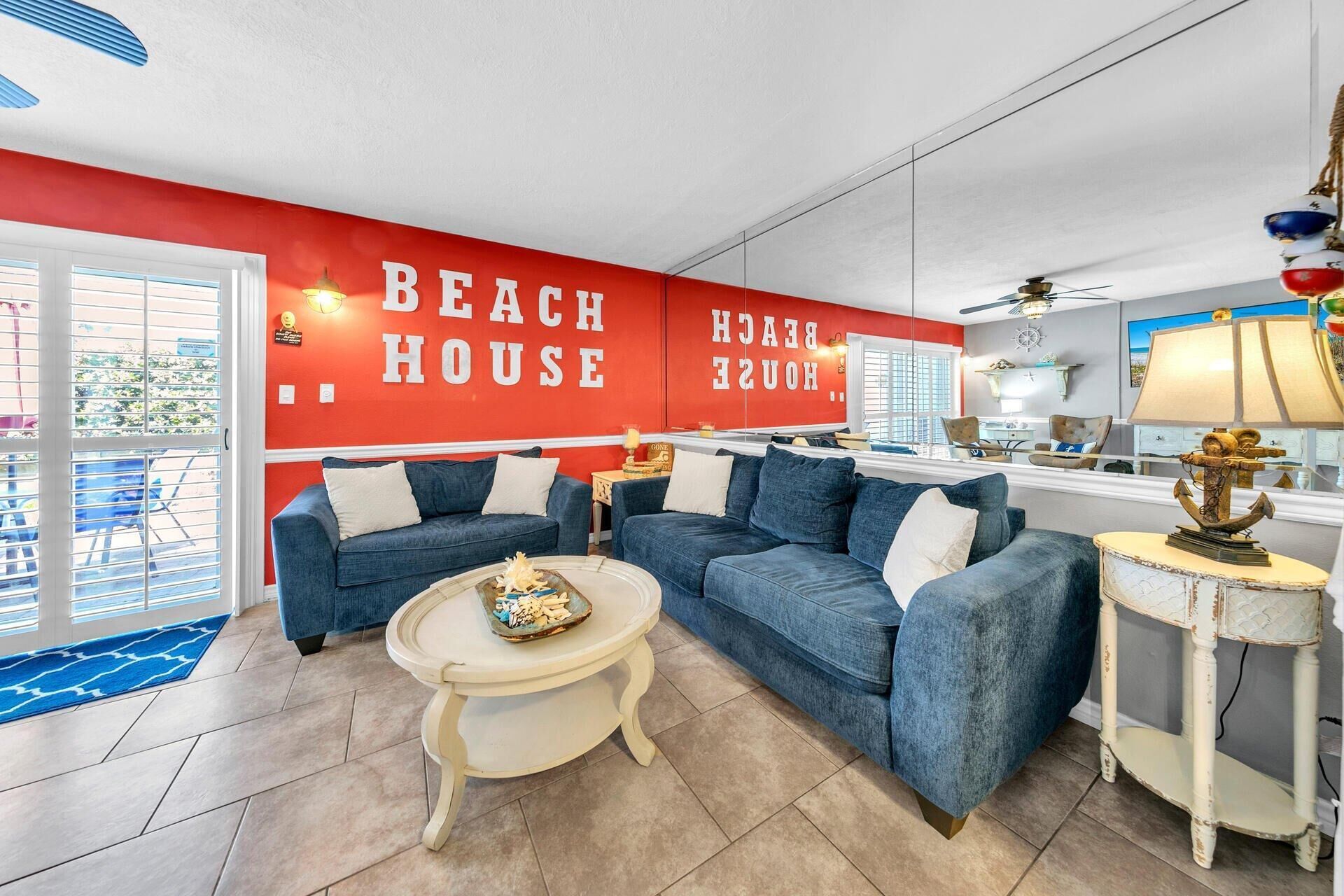 A living room with two blue sofas, a white coffee table, and a red wall with "BEACH HOUSE" text.