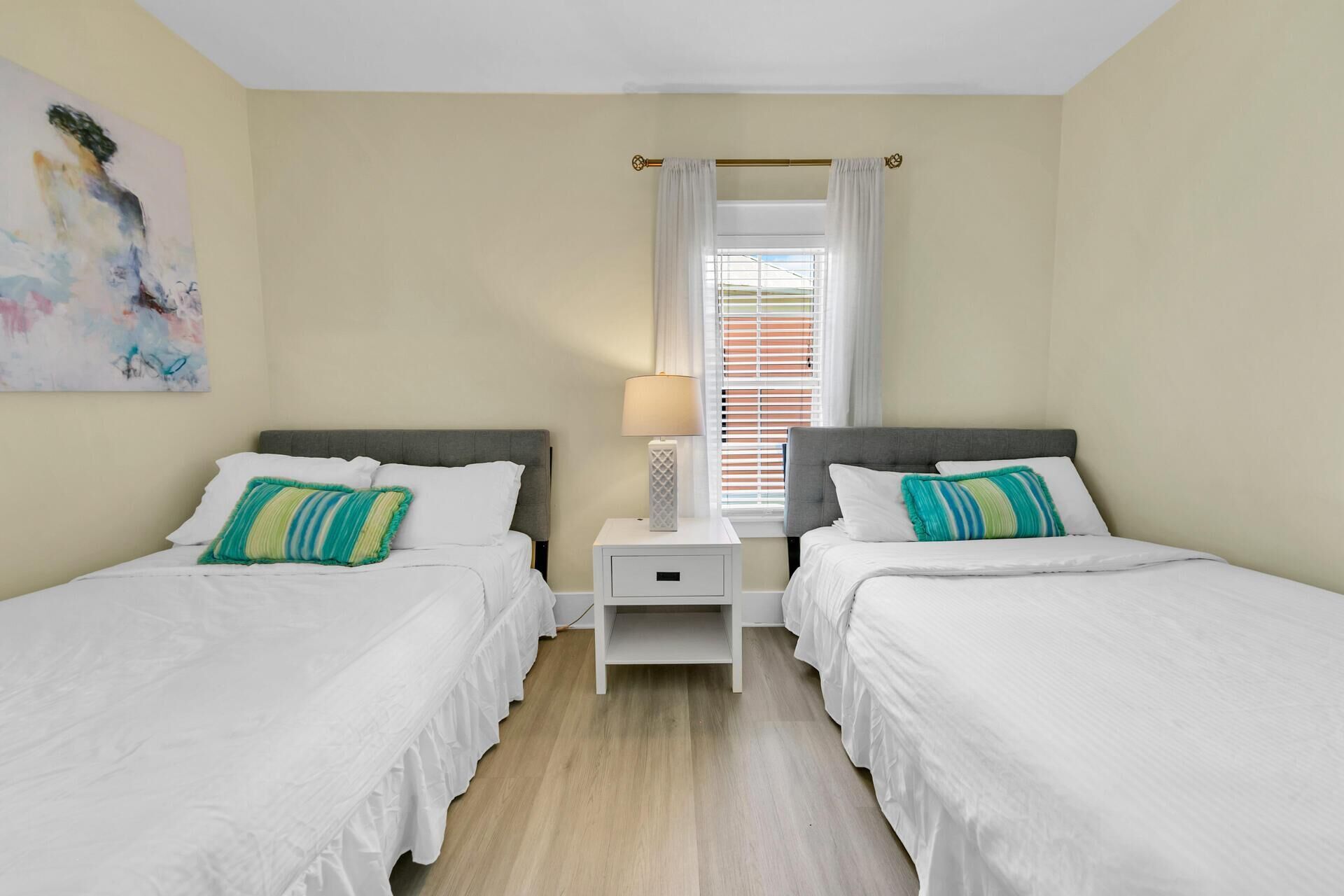 A hotel room with two single beds, a nightstand with a lamp, a window with blinds, and a painting on the wall.