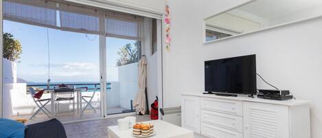 A modern living room with a large TV, a dining area with a table and chairs, and a balcony with a view of the sea.