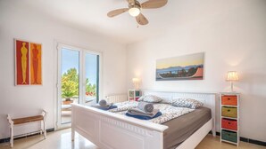A bedroom with a bed, a bench, a ceiling fan, a window, and a painting. - Pinomar by Interhome (Colònia Sant Pere)