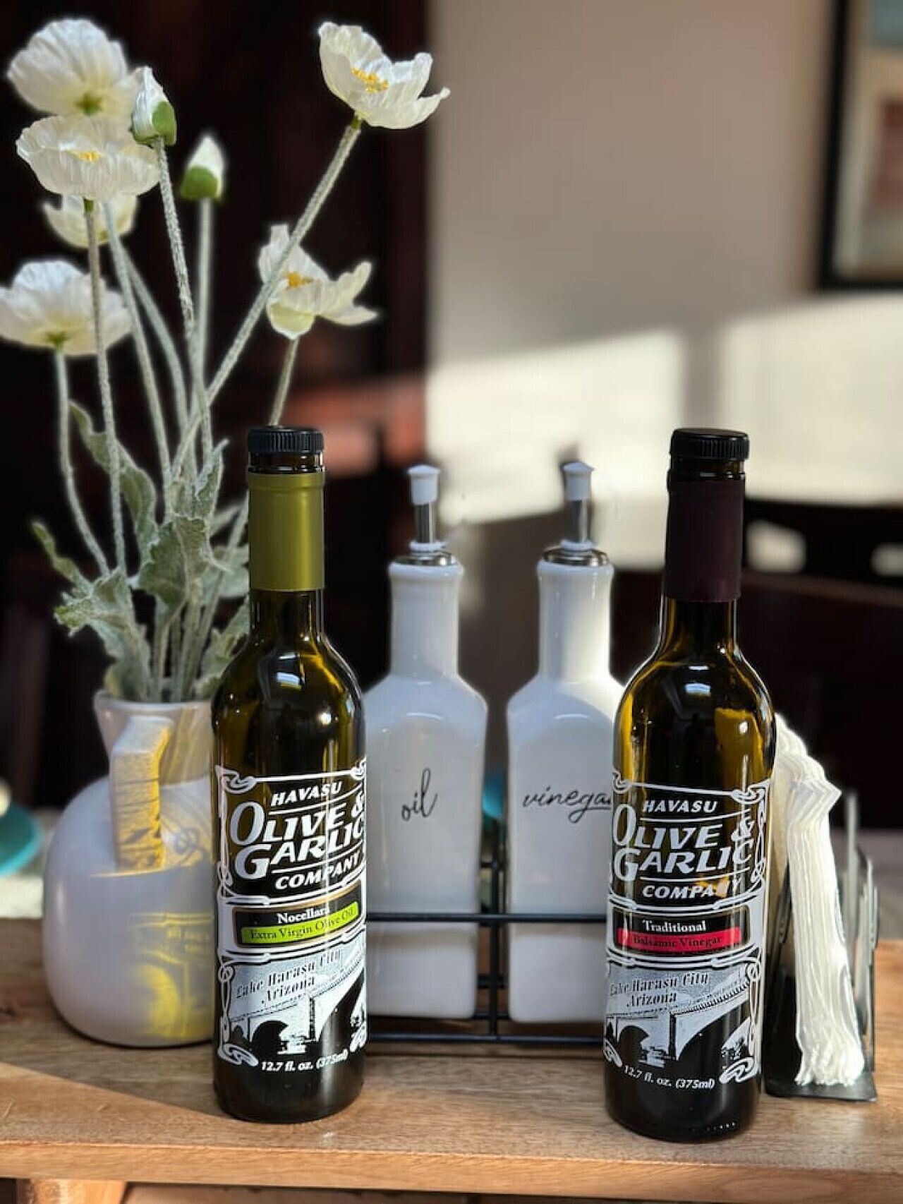 Two bottles of Havasu Olive & Garlic Company products, a white vase with flowers, and a clear bottle rack with additional items.