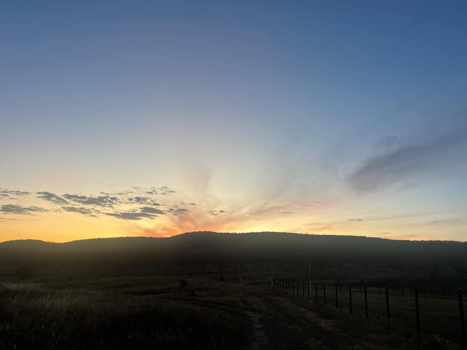 Paisaje rural con un camino de tierra, una cerca y un horizonte montañoso al atardecer.