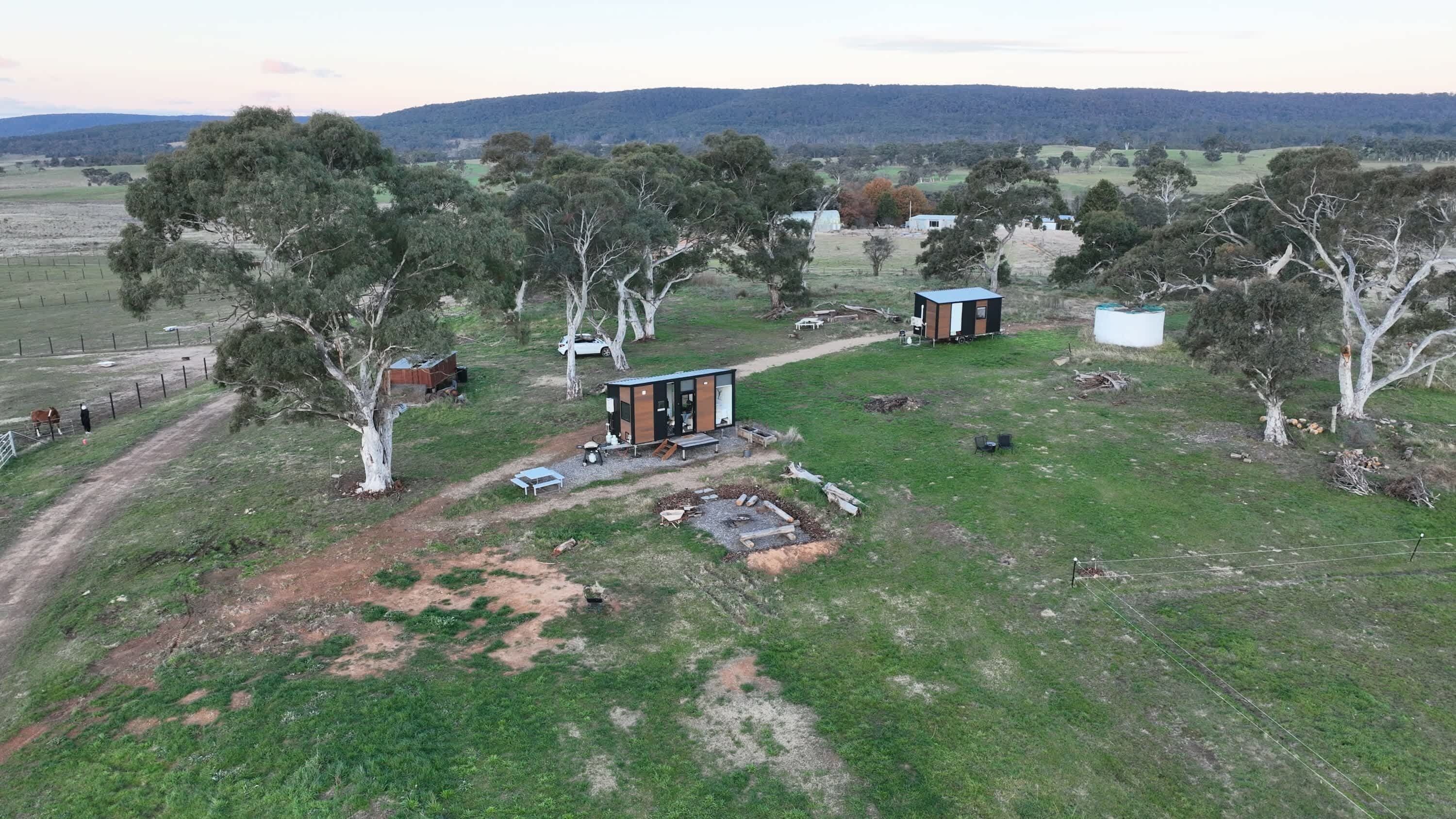 A rural property with a small cabin, a fenced paddock with a horse, and a hilly landscape in the background.