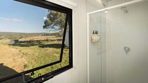 Et værelse med et vindue der kigger ud over en græsplæne, en hvid dør med brusebad og en lille hylde med toiletartikler.