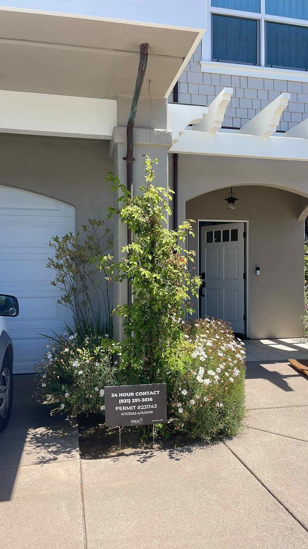 Edifício residencial com porta de garagem, entrada para carros e placa indicando informações de contato e licenças 24 horas.