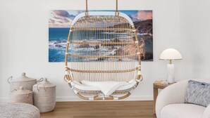 Una habitación con una silla colgante de ratán, una mesita de madera con una lámpara y un cuadro grande de un paisaje costero en la pared.