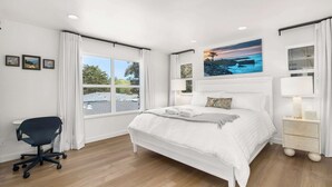 Un dormitorio con una cama grande, un escritorio y una silla. Hay tres cuadros en la pared, una ventana con cortinas y un cuadro sobre la cama.