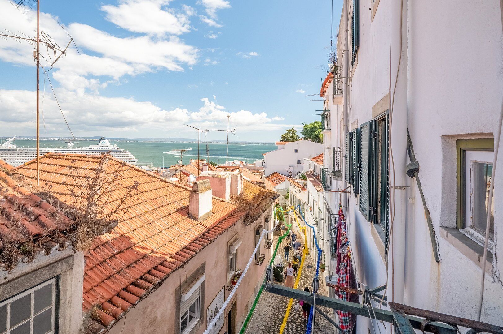 A narrow street with terracotta roofs, a white building with a balcony, and a view of the sea and a cruise ship in the distance.