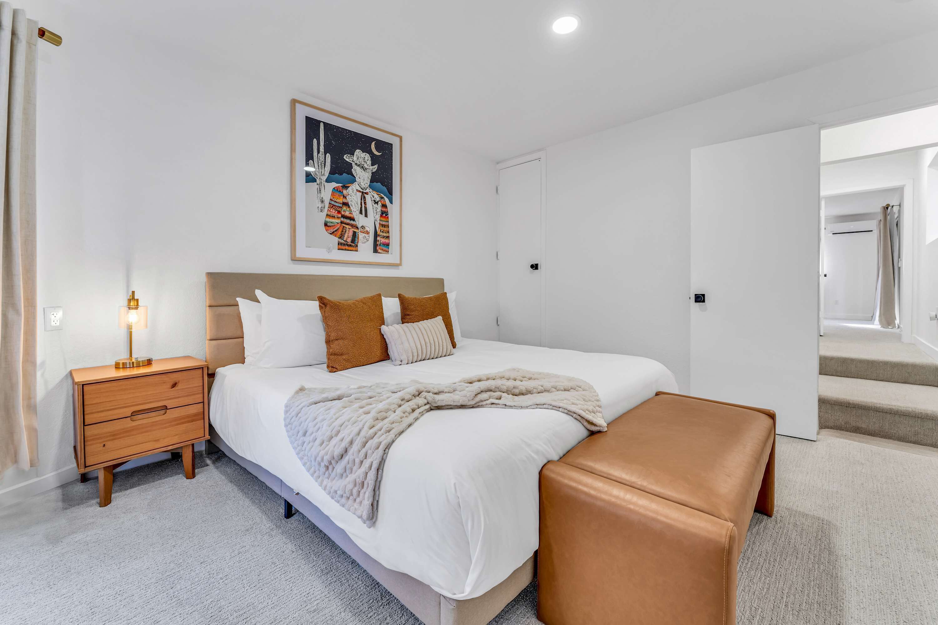 Une chambre à coucher moderne avec un grand lit, une table de chevet en bois, un banc et un tableau accroché au mur.