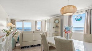 Ein modernes Wohnzimmer mit einer Couch, Esstisch und Stühlen, einem großen Fenster mit Meerblick und einem dekorativen Regal mit Gegenständen.