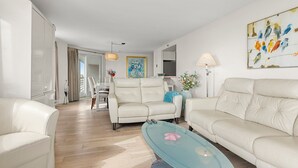 Ein modernes Wohnzimmer mit einem Glastisch, zwei weißen Sofas und einem Essbereich mit Tisch und Stühlen.