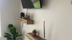 A modern living room with a flat-screen TV mounted on the wall, wooden floating shelves with decorative items, and a potted plant. - Don Cidales House (Pocosol)