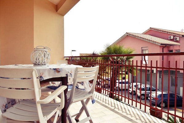 Un balcón con una mesa y sillas, un farol decorativo y vistas a una calle con coches aparcados y un edificio al fondo.