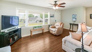 A living room with a flat-screen TV, two sofas, a ceiling fan, and large windows.