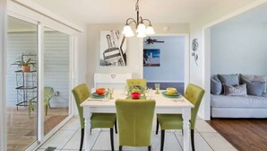 A dining area with a white table, green chairs, and a chandelier. There is a sofa and a wall with artwork in the background.