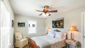 A bedroom with a wooden bed, two bedside tables with lamps, a ceiling fan, a window with blinds, and two wall-mounted pictures.