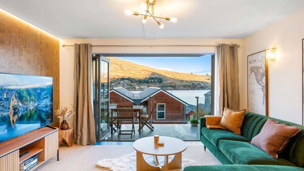 A modern living room with a television, a green sofa, a coffee table, and a view of a mountain and lake through large glass doors. - The Nugget-Free Parking-Direct Lake Access (Queenstown)